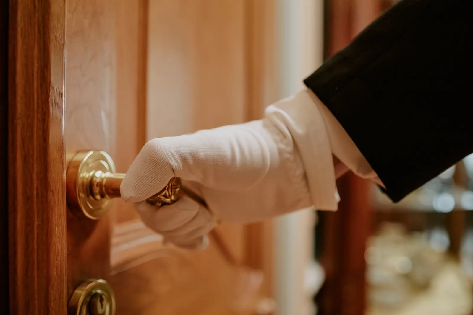 A white-gloved hand turning the brass handle of a polished wood door