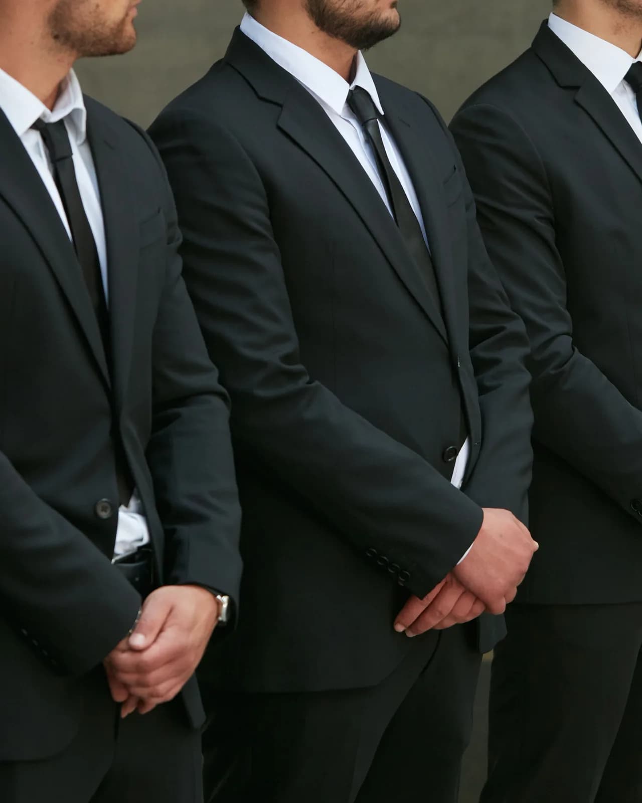 A row of men in dark suits and ties standing with hands clasped, cropped just below the eyes