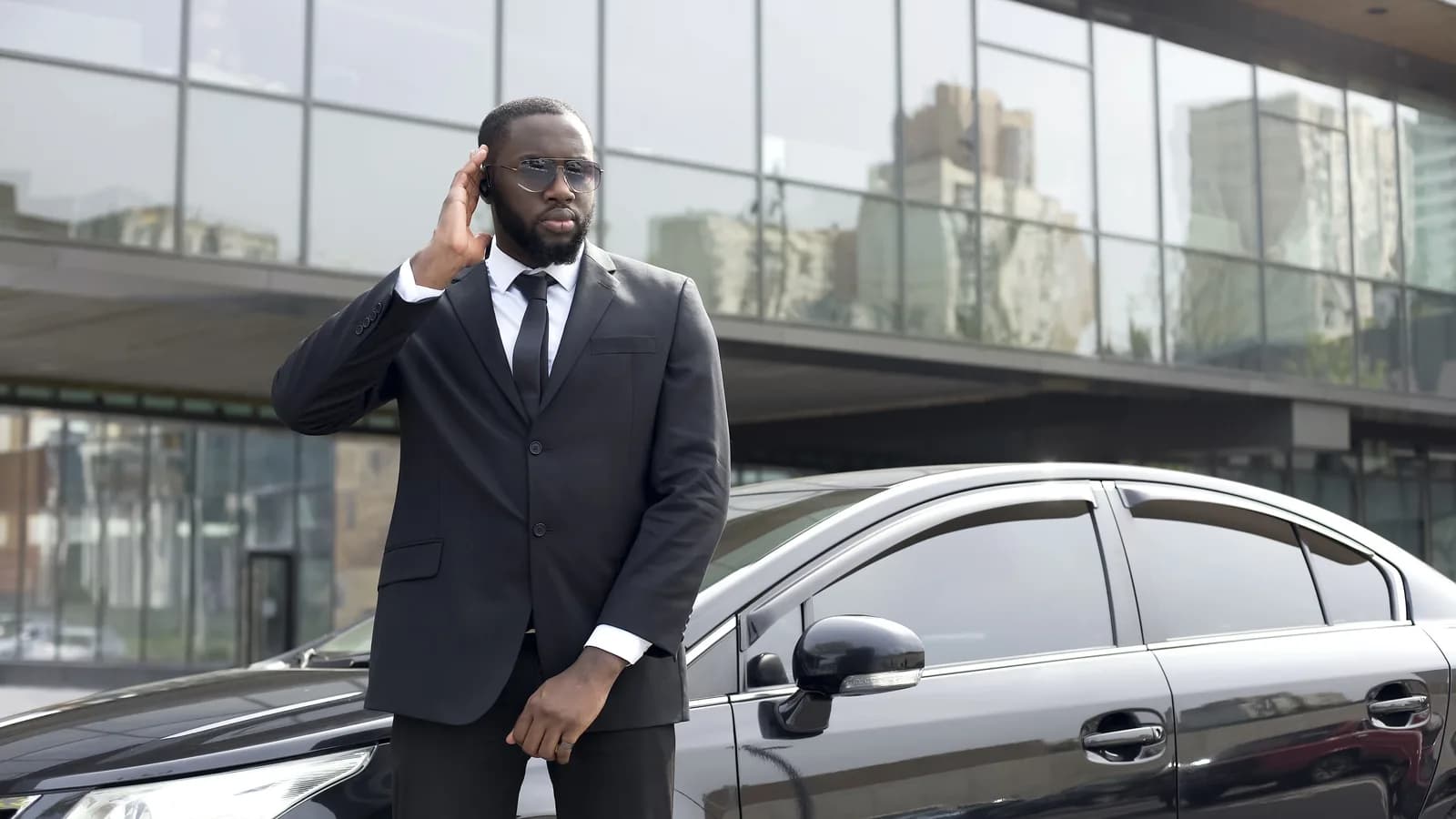 A man in a dark suit and sunglasses beside a black car, one hand to his earpiece