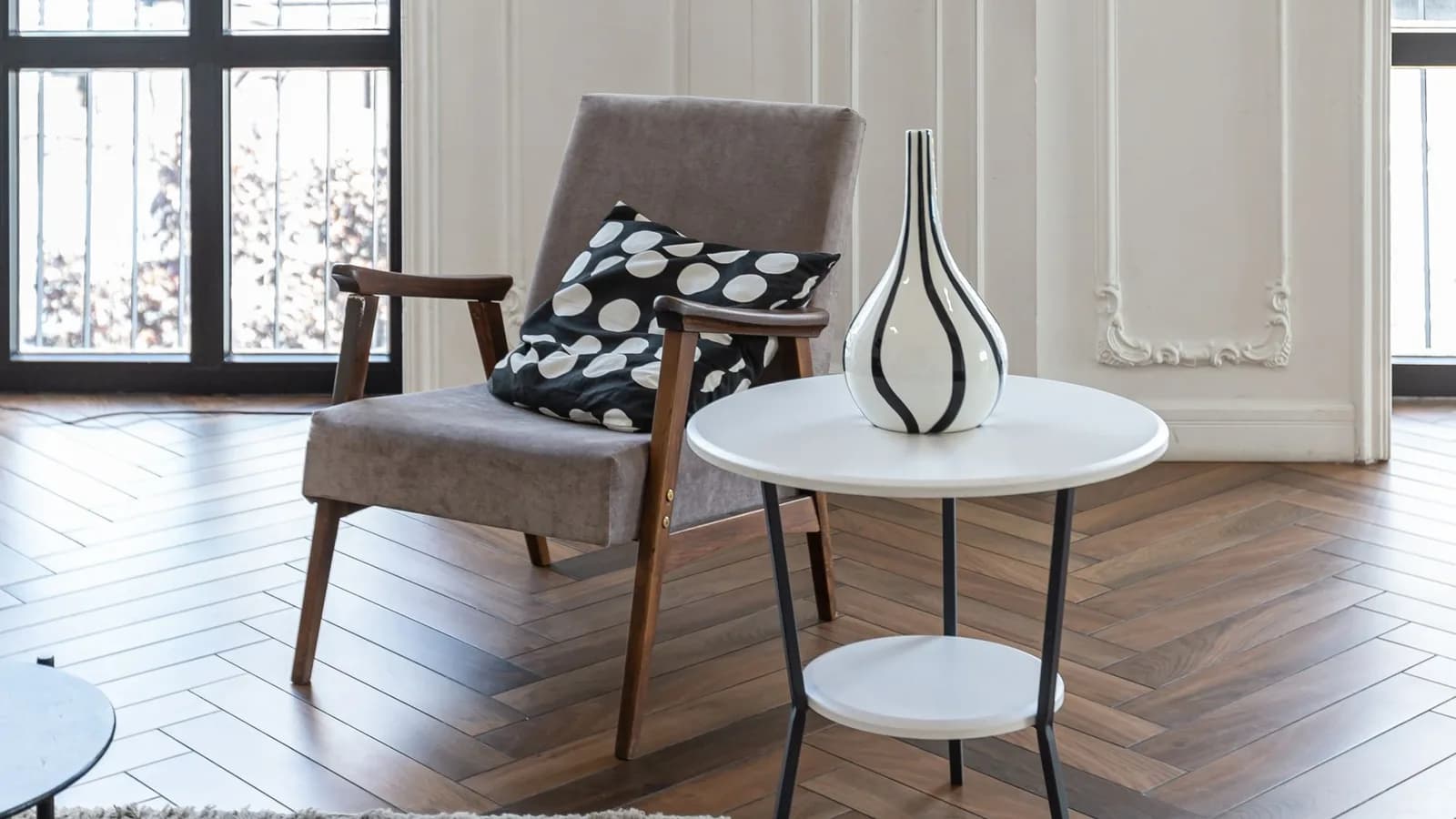 An armchair and a small round table with a vase on herringbone parquet, panelled white walls and tall windows behind