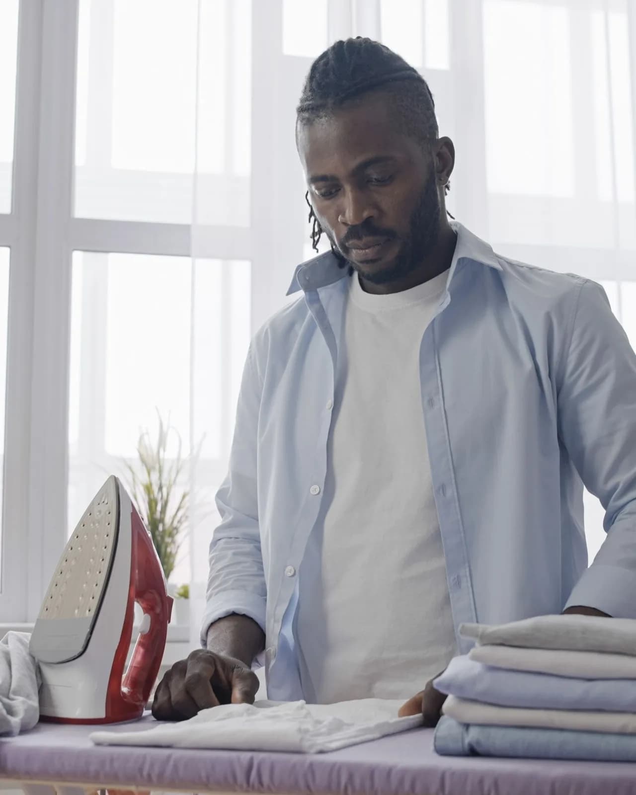 A man folding pressed shirts at an ironing board by a bright window, a red iron upright beside him