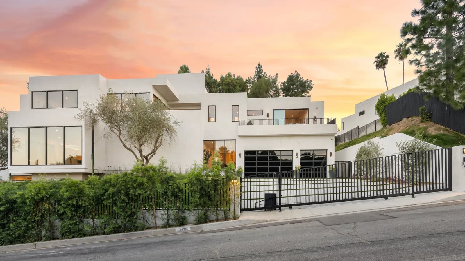 A modern white house with olive trees and a clipped hedge behind a low black gate at sunset