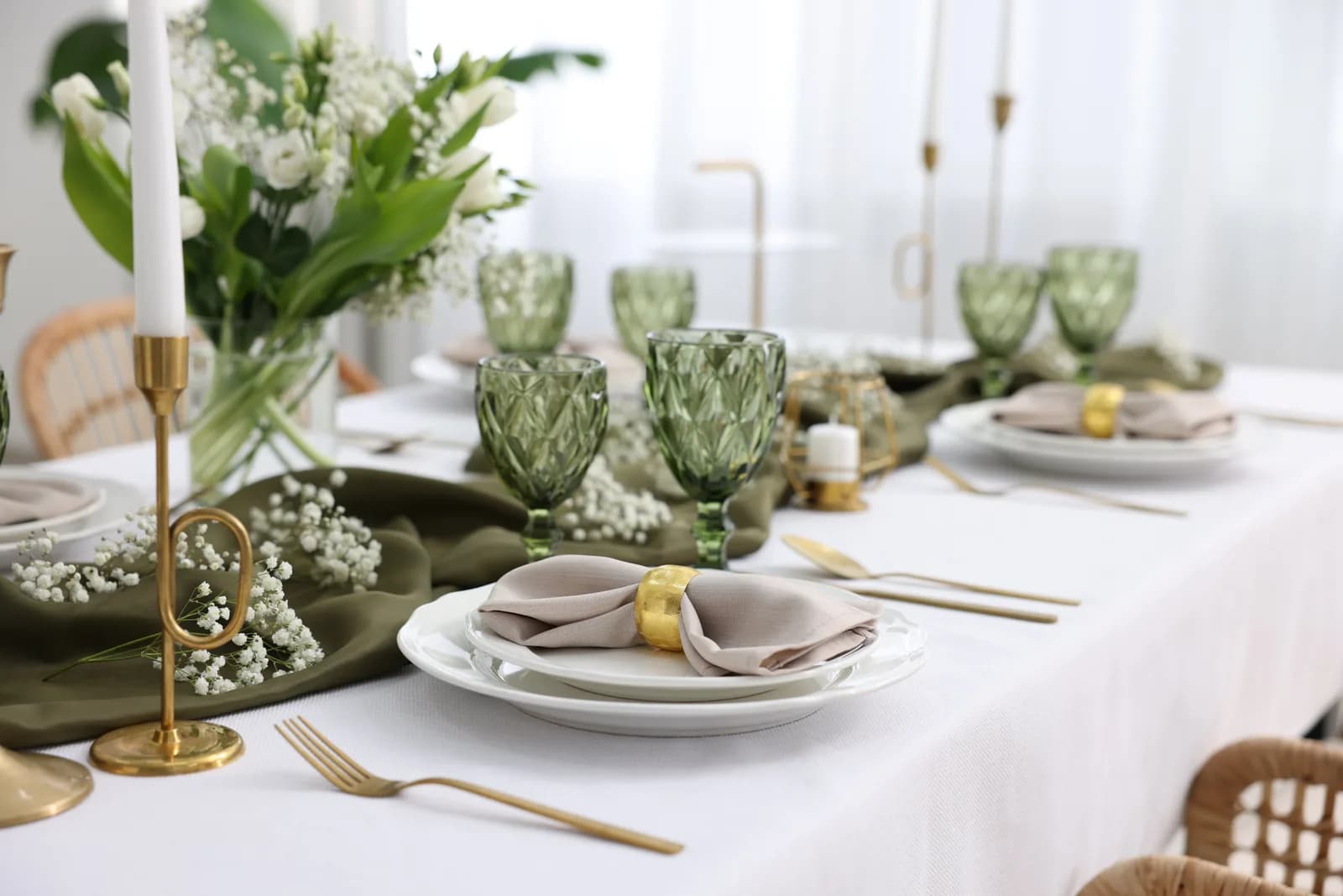 A table laid with green glassware, gold cutlery and white flowers on white linen