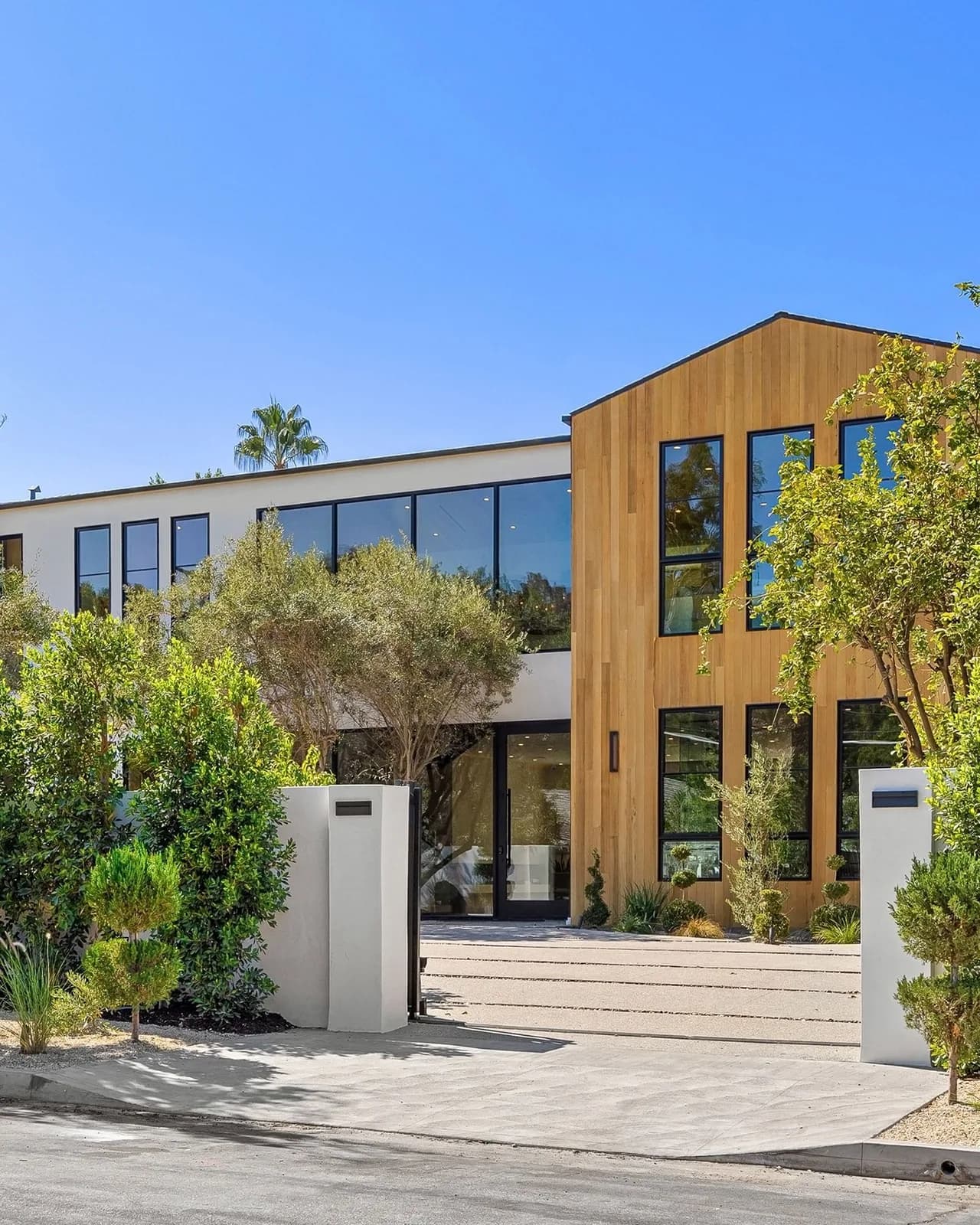 A two storey house in white render, timber and glass behind clipped hedges and a paved drive