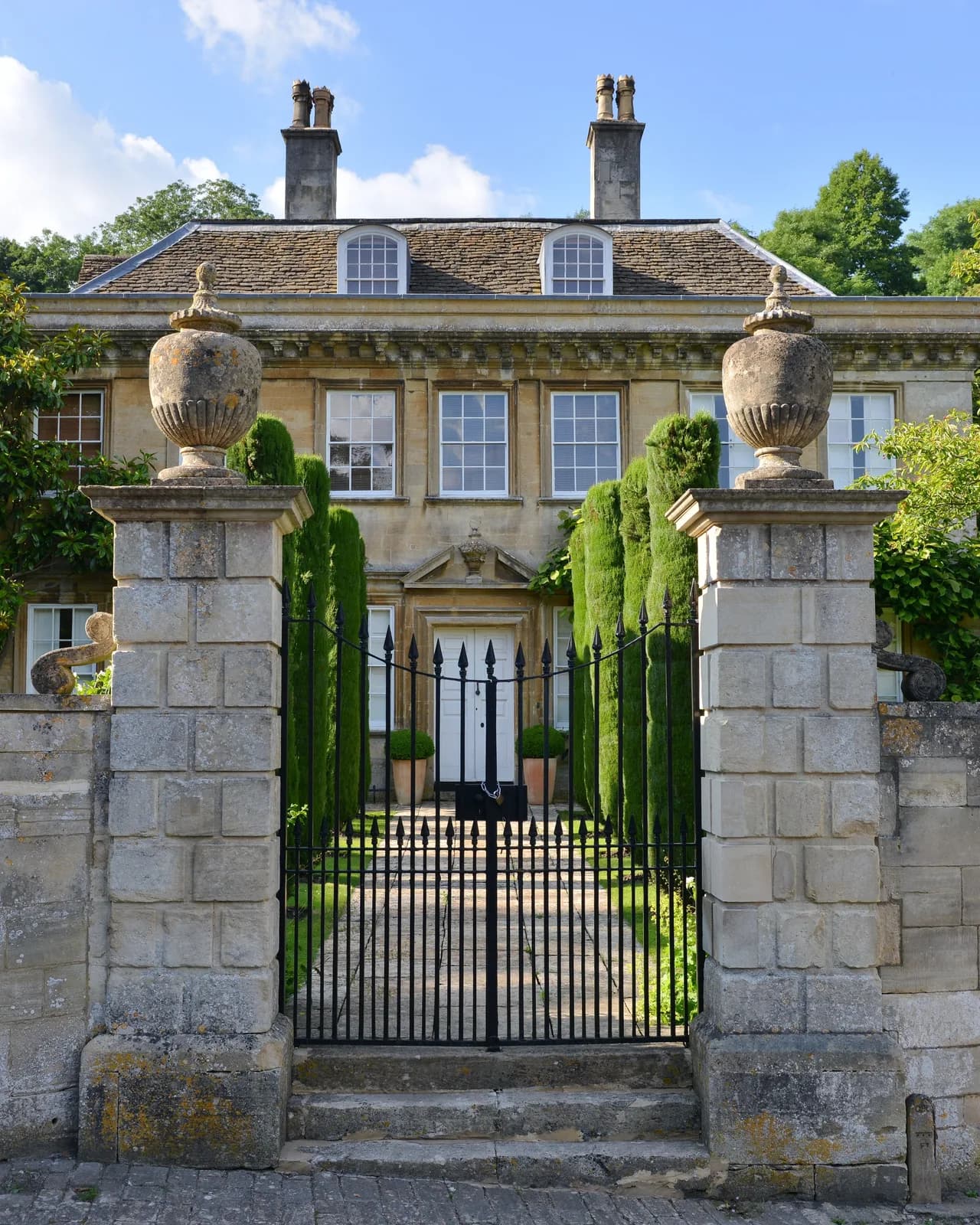 An English country house of pale stone behind iron gates set between urn-topped piers