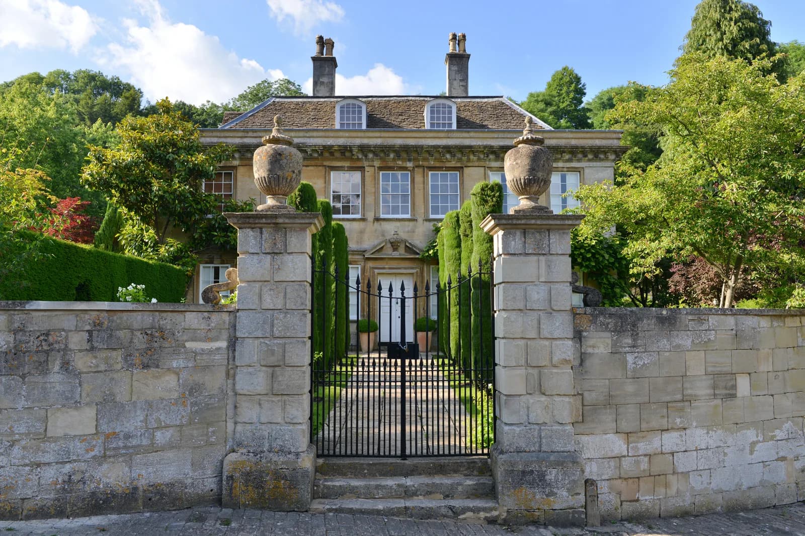 A stone country house seen through iron gates flanked by urn-topped piers