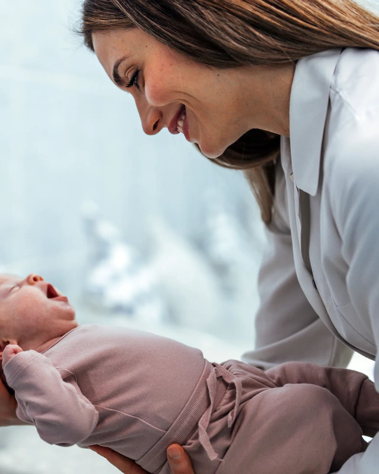 A woman leaning over a yawning newborn in a pink knit sleepsuit, held in her hands