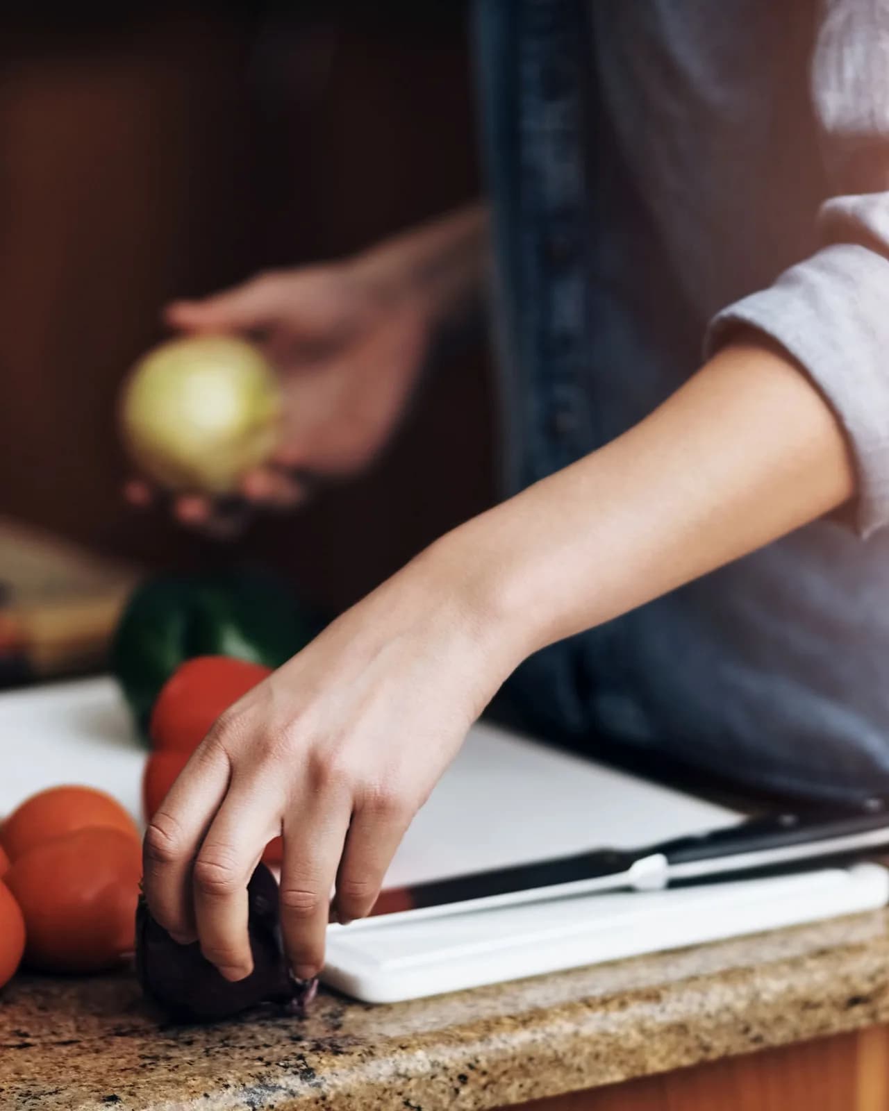 A cook's hands at a stone counter with an onion, tomatoes and a chopping board